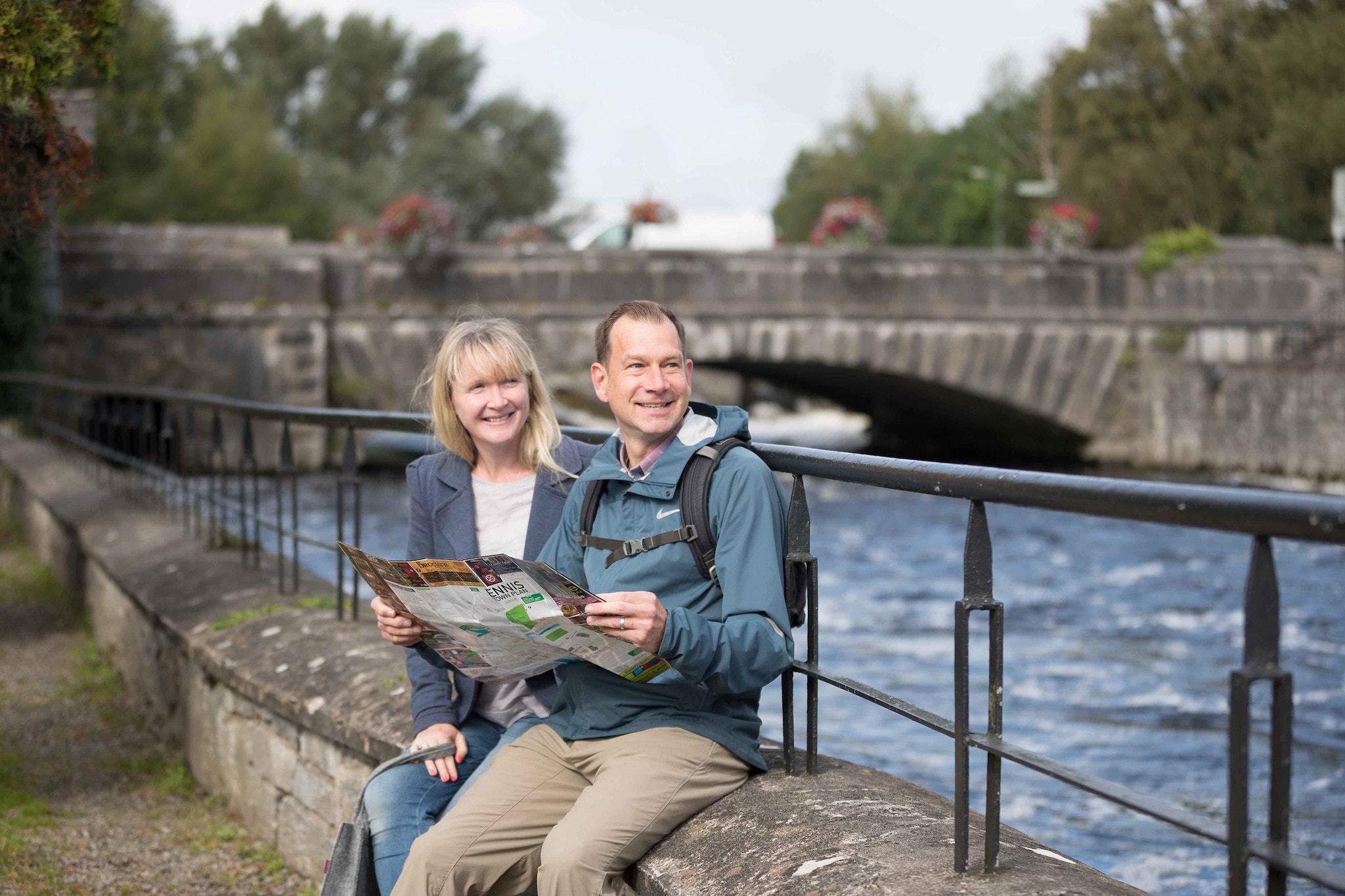 A smiling couple sits on a low stone wall beside a river in Ennis, Ireland, holding an open "Ennis Town Plan" tourist map, with a stone arch bridge decorated with flower baskets behind them.